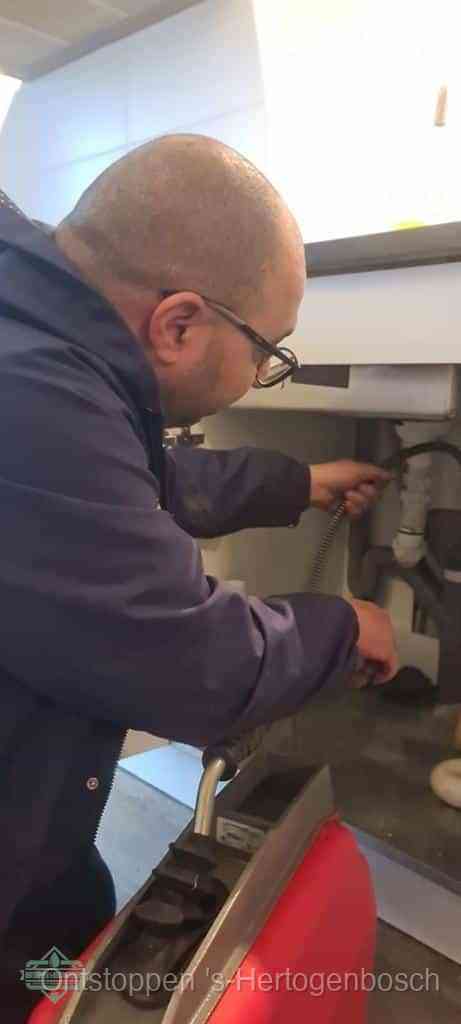 Een monteur buigt zich onder een wastafel en voert een ontstoppingsveer in om de bron van rioollucht uit de afvoer in s-Hertogenbosch op te sporen