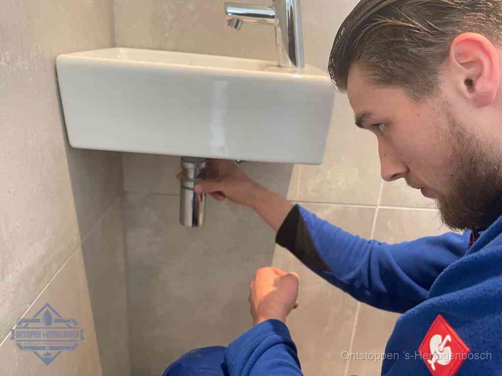 Een monteur draait met de hand de zwanenhalssifon onder een fonteinwasbak in de toiletruimte los om de bron van rioollucht in s-Hertogenbosch te vinden