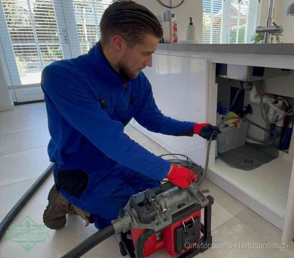 Een monteur met rode handschoenen knielt in een keuken en voert een elektrische ontstoppingsveer in tijdens het professioneel afvoer ontstoppen in s-Hertogenbosch